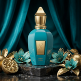 A turquoise and gold perfume bottle labeled 'ERRBA PURA' stands on a dark marble pedestal, surrounded by decorative gold flowers, a pomegranate, and a deep teal draped fabric background.