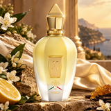 A faceted glass perfume bottle with a metallic gold cap and gold detailing, labeled NAXOS, set on a stone surface with white flowers, a lemon slice, and draped cream fabric. A scenic coastal background is visible behind it.