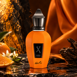 An orange perfume bottle with a black cap and a black label reading 'DURAN DURAN' and 'Neo Fo' is centered on a dark reflective surface. The background features draped orange fabric, a sliced orange, white flowers, and dark wood driftwood.