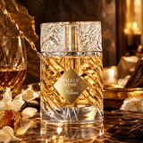 Angels Share Kilian perfume bottle made of clear cut crystal with a gold label, set on a reflective marble surface with warm golden lighting, white flowers, and a glass of amber liquid in the background.