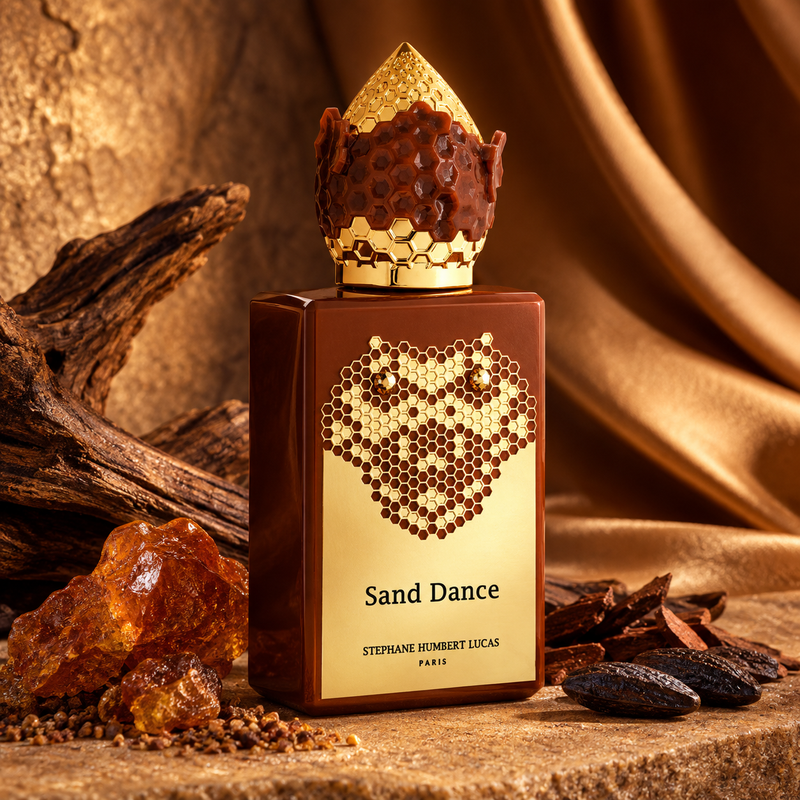 A perfume bottle labeled 'Sand Dance' by Stephane Houbert Lucas Paris, styled with amber resin, spices, and draped gold fabric in a warm, luxurious setting.