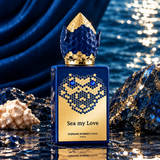 A bottle of Stephanie Hembert Lucas 'Sea my Love' perfume stands on a reflective wet surface with blue draped fabric, rocks, and a shell around it. The bottle is deep blue with a gold honeycomb-patterned face and cap, and the text 'STEPHANIE HEMBERT LUCAS PARIS' is visible on the front.