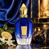 A luxury perfume bottle with a deep cobalt blue body and a metallic gold cap, styled on a reflective surface with a blue satin drape, a lemon slice, white flowers, and a dark textured stone in the background. The bottle is labeled 'XERJOFF' and 'TORINO 21'.