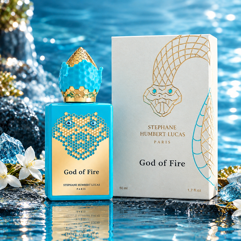 A perfume bottle and its box for Stephane Humbert Lucas God of Fire, styled on a reflective blue water surface with rocks and white flowers. The bottle is bright blue with gold and white graphic details, and the box is white with gold snake artwork and blue accents.