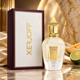 Xerjoff Shooting Star Lua perfume bottle with a gold cap and clear glass body, shown next to its dark purple and white box on a reflective surface with draped fabric and citrus slices in the background.
