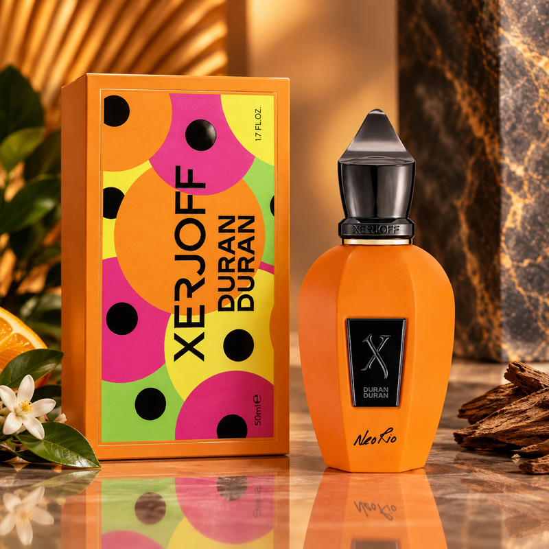 Xerjoff Duran Duran perfume bottle and its orange packaging box, styled on a reflective marble surface with decorative elements including a white flower, a slice of orange, and wood pieces.
