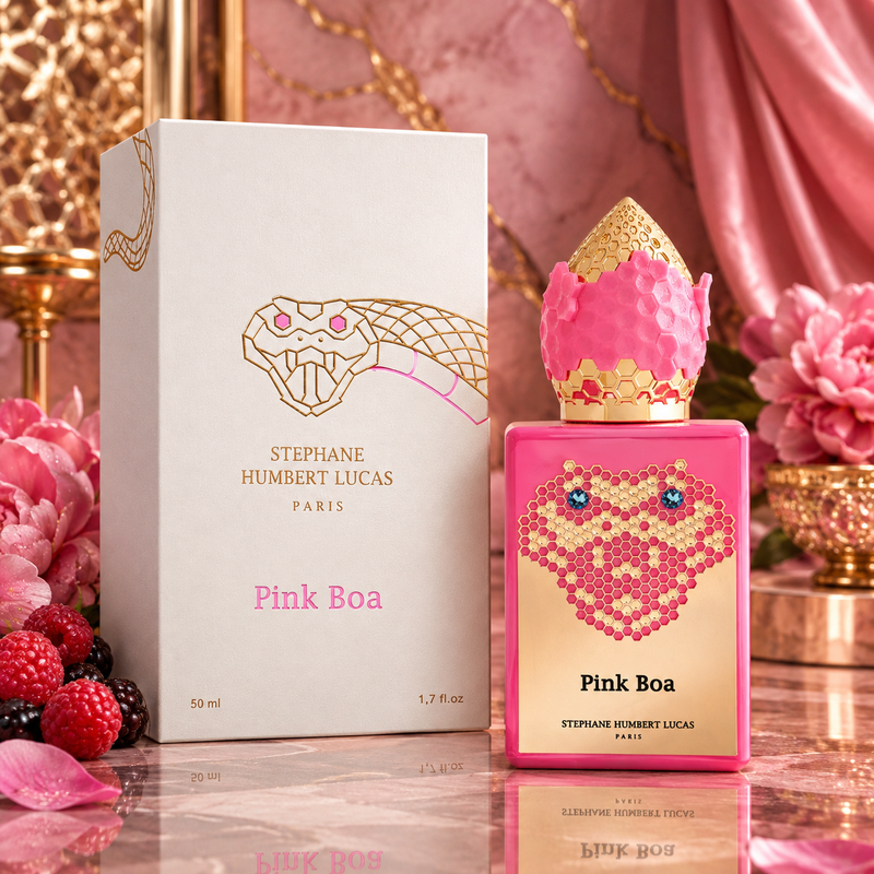 Pink Boa perfume by Stephane Humbert Lucas, showing the pink and gold bottle next to its white and gold box, styled with pink flowers and berries on a reflective surface.