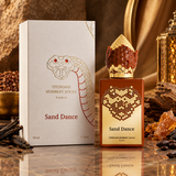 A perfume bottle and its box for Stephane Humbert Lucas Sand Dance, styled with amber resin, dried herbs, and gold-toned decorative props on a reflective surface.