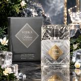 Vodka Kilian On the Rocks eau de parfum 50ml bottle with a faceted clear glass cap and a gold label, shown next to its dark gray box. The scene is styled with ice cubes, pine branches, white flowers, and a reflective dark surface.