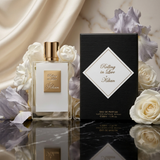 A perfume bottle with a gold cap and gold label stands beside its black packaging box with a white diamond label reading 'Rolling in Love Kilian.' The bottle and box are styled with white and pale lavender flowers on a reflective dark marble surface, with draped satin fabric in the background.