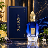Xerjoff Torino 21 perfume bottle and its navy blue box, styled on a reflective surface with blue fabric, flowers, and dark berries.