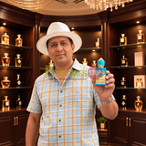 A man wearing a white hat and a light blue plaid shirt holds up a small blue perfume bottle labeled 'Sultan of Oman' in front of a dark wood display cabinet filled with other perfume bottles.