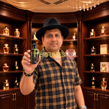 A man wearing a black hat and a brown and black plaid shirt holds up a perfume bottle labeled 'X' in front of a background of wooden shelves filled with other perfume bottles.