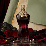 "A Jean Paul Gaultier La Belle Le Parfum perfume bottle with a gold-toned corset design and dark red liquid, set against an elegant background with red roses and soft lighting."