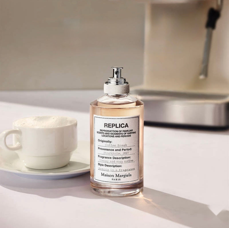 "A Maison Margiela Replica Coffee Break perfume bottle with a cream-colored label, set against a cozy coffee shop background with a cup of coffee and soft natural lighting."