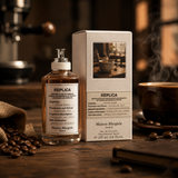 "A bottle of Maison Margiela Replica Coffee Break Eau de Toilette with its matching packaging box, set against a cozy coffee shop background with coffee beans and warm lighting."