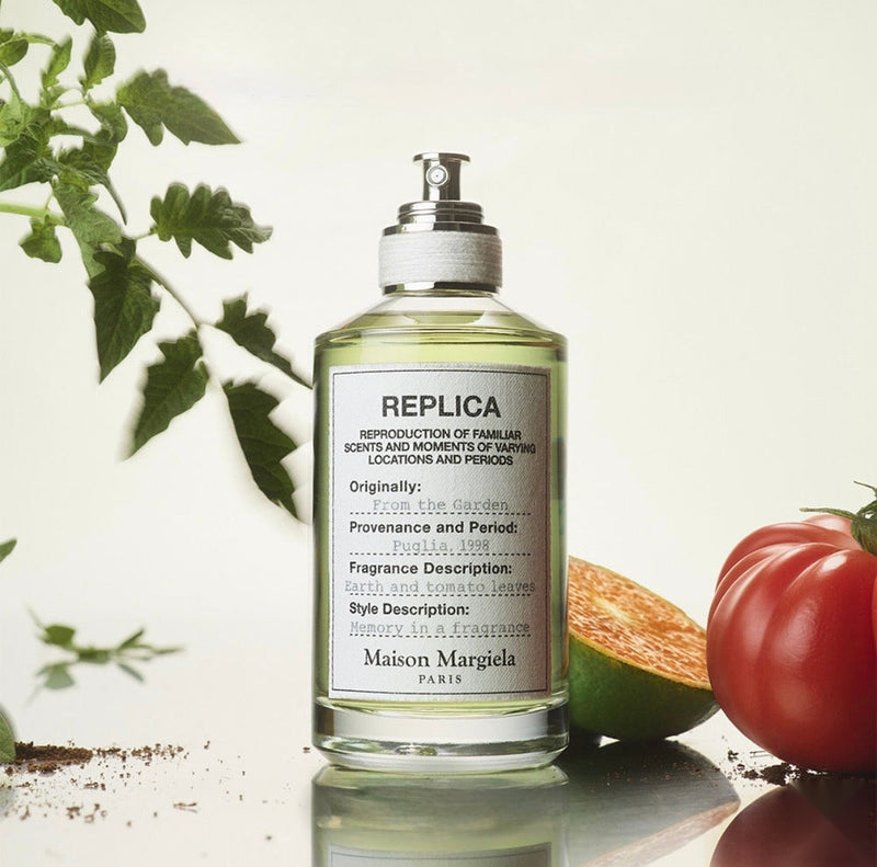 "A clear perfume bottle labeled 'Replica From The Garden' by Maison Margiela, featuring a white label and silver cap, set against a bright garden background with green foliage, tomato leaves, and soft natural lighting."