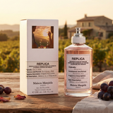 "A bottle of Maison Margiela Replica On A Date Eau de Toilette with its matching white packaging box, featuring a vineyard-inspired label, set against a warm countryside background with soft golden lighting."