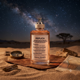 A bottle of Maison Margiela Replica Under the Stars fragrance is displayed in a desert setting with a night sky, stars, and Milky Way in the background. The bottle is placed on a leather pad with a compass, small bottle, and map nearby.