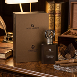 Swiss Arabian Tobacco 01 Extrait de Parfum bottle and its matching brown presentation box, styled on a wooden desk with cigars, a cigar ashtray, dried coconut, and books in the background.