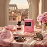 "A pink perfume bottle labeled 'Born in Roma Donna' by Valentino, displayed against a warm architectural background with pink roses and soft golden lighting."