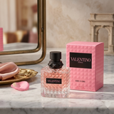 "A bottle of Valentino Born in Roma Donna Eau de Parfum with its matching pink packaging box, featuring floral decorations, set against a warm, elegant background with soft lighting."