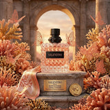 "A pink perfume bottle labeled 'Born in Roma Donna Coral Fantasy' by Valentino, displayed against a warm, elegant background with pink roses, flowers, and soft golden lighting."