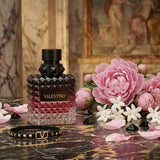 "A dark pink perfume bottle labeled 'Born in Roma Donna Intense' by Valentino, displayed against a floral background with pink roses and warm golden lighting."