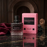 "A bottle of Valentino Born in Roma Donna Intense Eau de Parfum with its matching pink packaging box, featuring a dark floral design, set against a dramatic background with warm lighting."